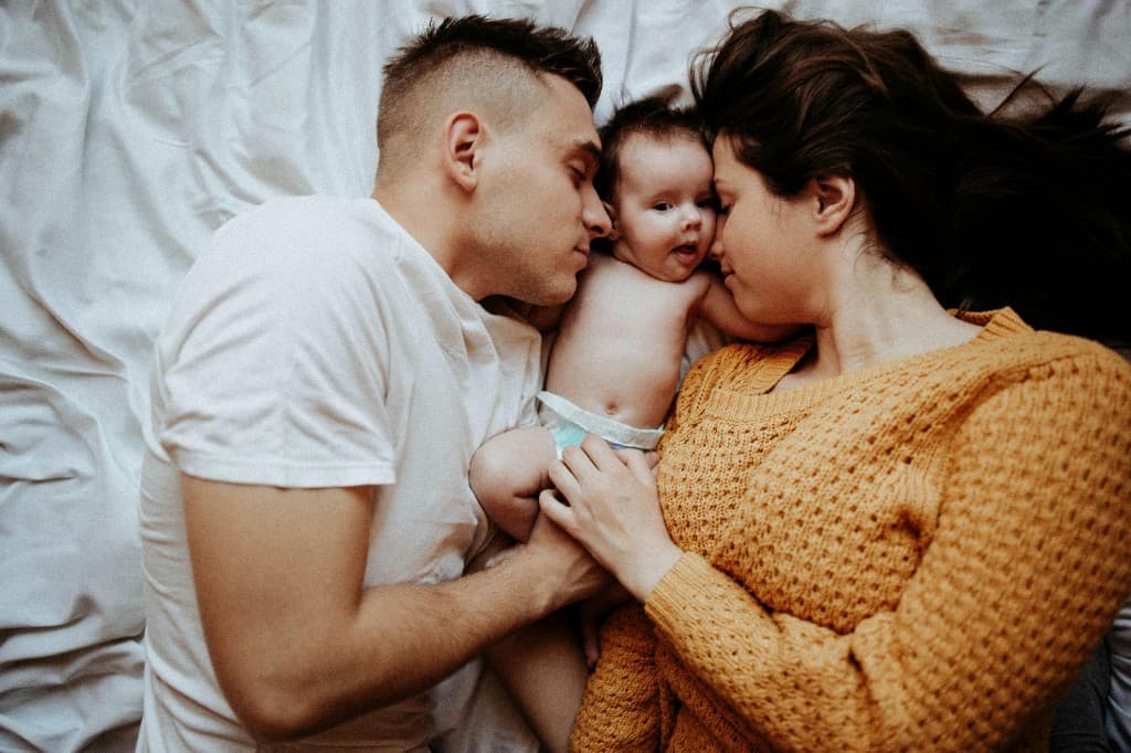 Parents endormis dans un lit avec la tête vers leur bébé éveillé entre eux, draps blancs, lumière douce